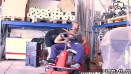 Blonde reife Chefin in schwarzem Kleid reitet einen Arbeiter auf einem roten Hubwagen im Lagerhaus